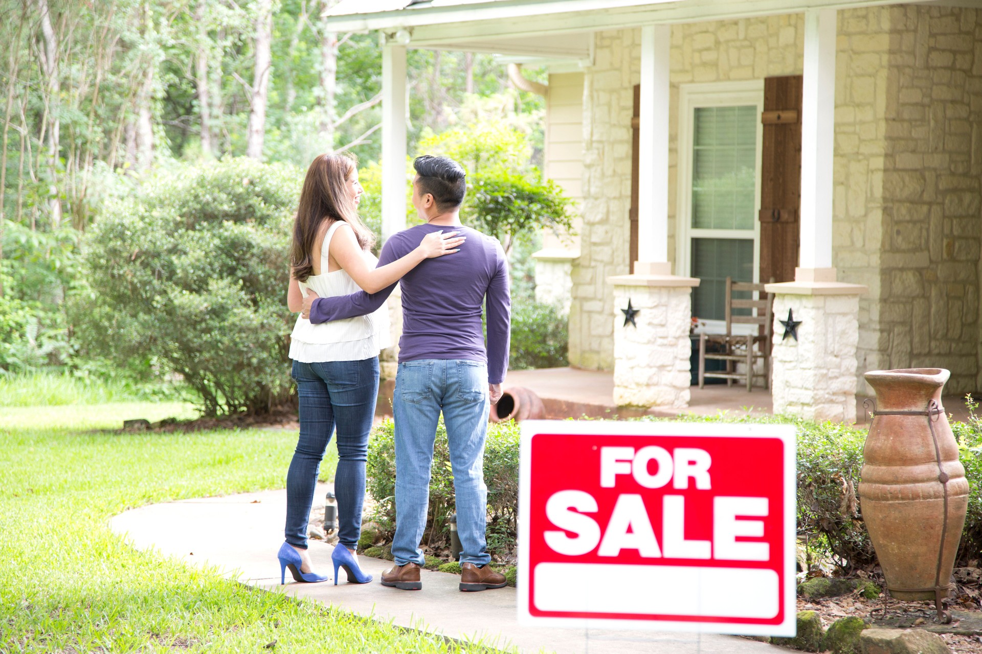 Mid adult Asian couple are considering purchasing a new home.  For sale sign in foreground.